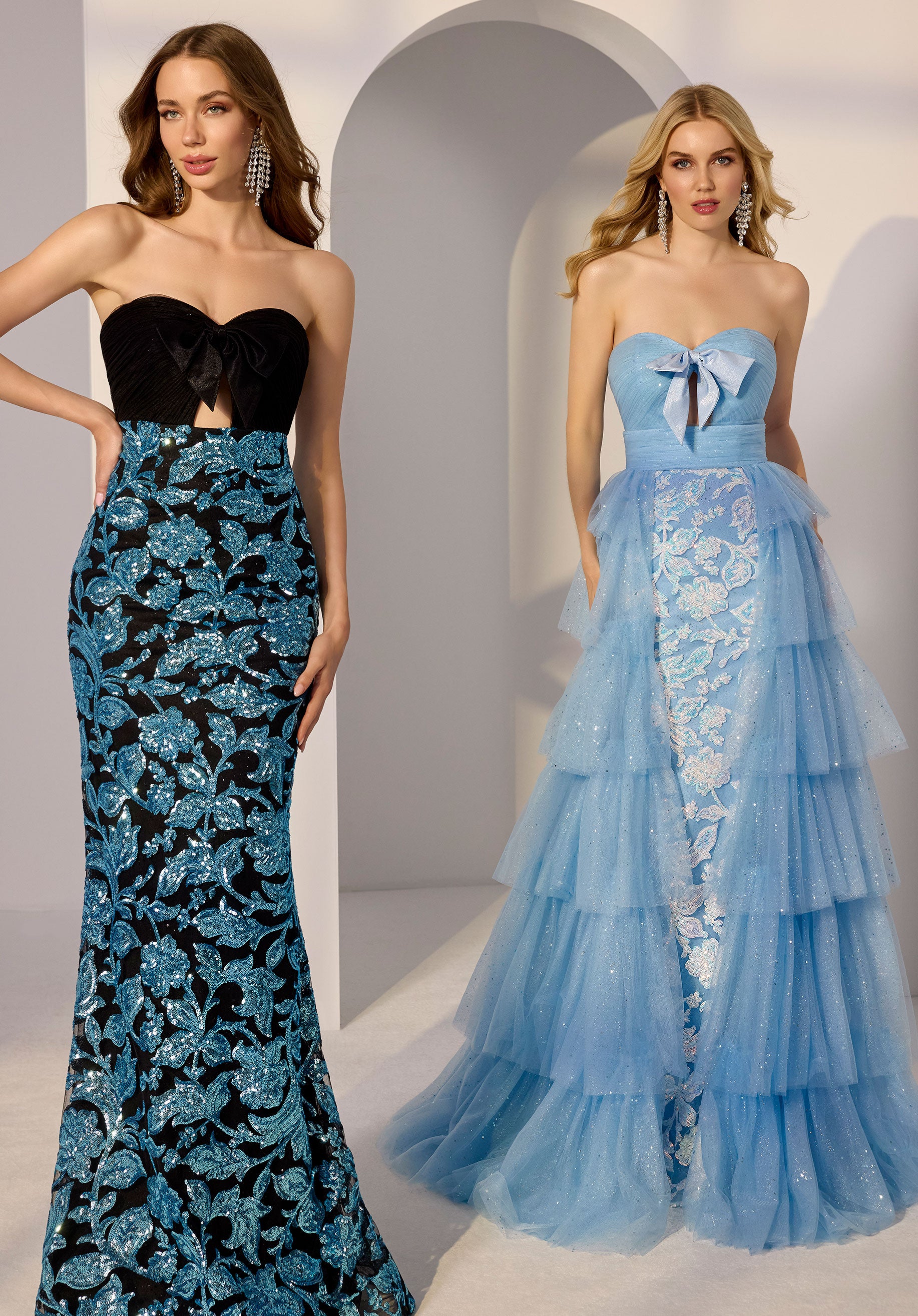 Two women stand side by side; one wears the Morilee 49275 long prom dress, featuring a strapless light blue design with lace detailing, a bow accent, and a detachable overskirt. The other wears a strapless black and blue floral gown.